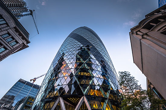 Vista desde abajo del icónico edificio Gherkin en Londres, con su distintiva arquitectura de vidrio y acero, rodeado de otros rascacielos y edificios modernos al atardecer.