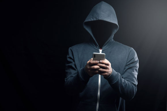 Persona con una sudadera con capucha, con el rostro oculto en la sombra, usando un teléfono inteligente en un fondo oscuro. La imagen simboliza el anonimato y la ciberseguridad o actividades de hacking