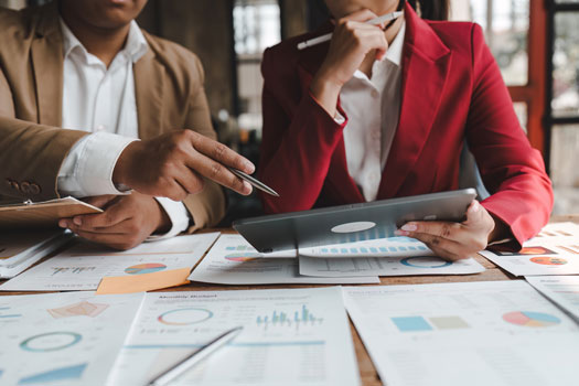 Dos profesionales de negocios revisando documentos y gráficos financieros en una mesa, mientras utilizan una tableta. Ambos están vestidos con trajes formales, uno en beige y el otro en rojo, en una reunión o entorno de trabajo colaborativo