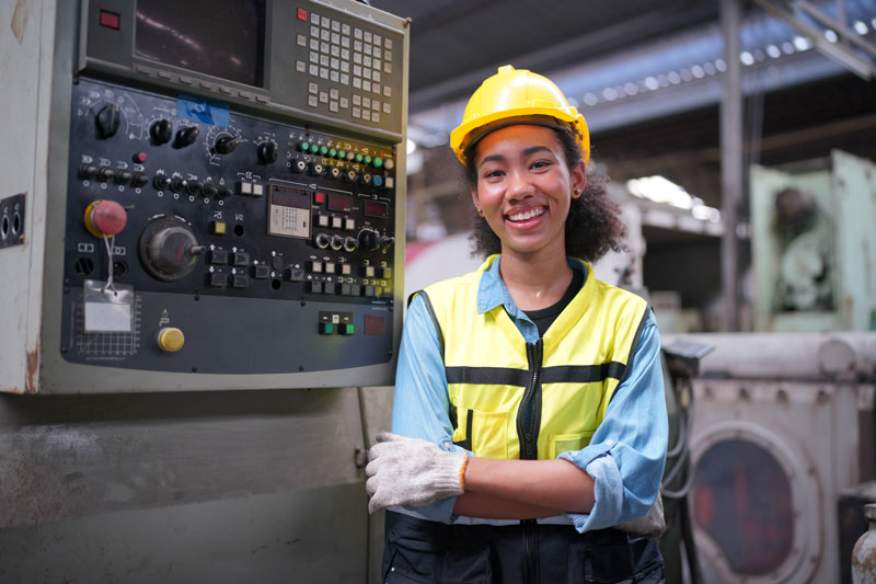Trabajadora de una fábrica, vestida con equipo de seguridad, incluyendo casco amarillo y chaleco reflectante, sonriendo frente a una máquina de control industrial en un entorno de manufactura.